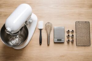An overhead view of a white stand mixer on pale oak with a spatula, balloon whisk, digital scale, six piping nozzles and a cooling rack arranged in descending size.